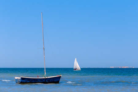 Photo of calm blue sea on a sunny day, where the foreground is a beautiful small sailboat, and in the background a sailboat with sails.It is seen in distant city.の写真素材