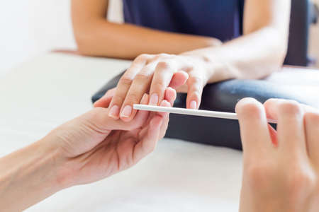 Manicurist performing a professional manicure in a beauty salon shaping and filing a clients fingernails with a nail file, close up view of their handsの写真素材