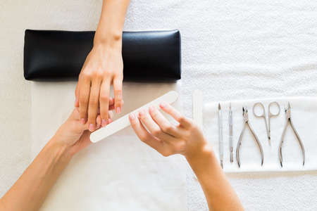 Lady receiving a manicure in a salon having her fingernails filed by a professional manicurist, view from above of their hands and toolsの写真素材
