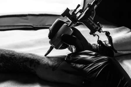 Close up picture of tattooist make a tattoo on clients arm in bw. Master works on the desk in black sterile gloves and hold a tattoo machine.の写真素材