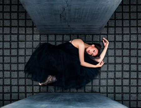 ballerina in black ballet tutu lainging lie on left side on gray tiles between slabs of concreteの写真素材