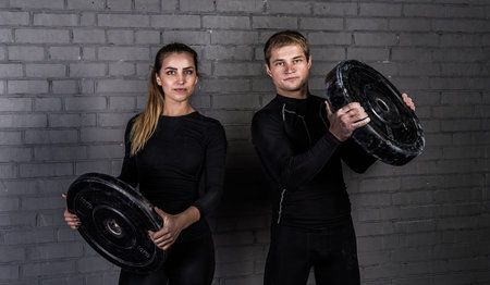 Couple of sporty man and woman holding barbells in hands and smiling. love sports near gray wall