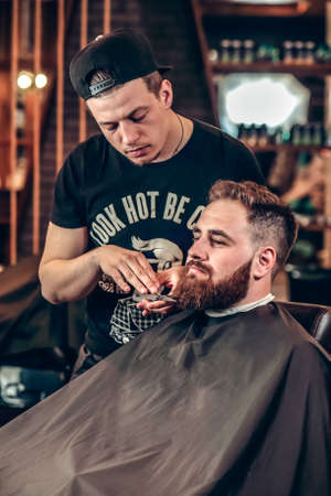 Young handsome barber making beard grooming of attractive bearded man in barbershop. Grooming of beard using professional scissors in barber shop. Bearded hipster in a barber shopの写真素材