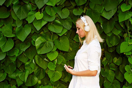 Female student girl outside in park listening to music on headphones. Blond woman having fun on a background of green leaves.の写真素材