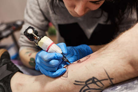 Tattoo artist is making tattoo. The process of creating a tattoo on the mans leg, closeup view. Machine for tattoos during work closeupの写真素材