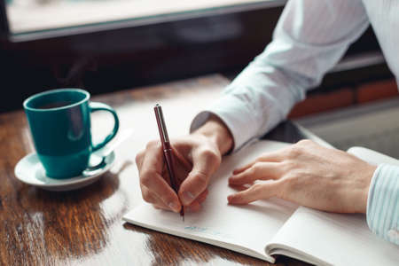 Close up of hands of a businessman writing in a notebook and a cup of coffee. A freelance man is working in a cafe. Coffee break.の写真素材