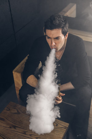 Vaping man holding a mod. A cloud of vapor. Black background. Smoking electronic cigarette Vape...