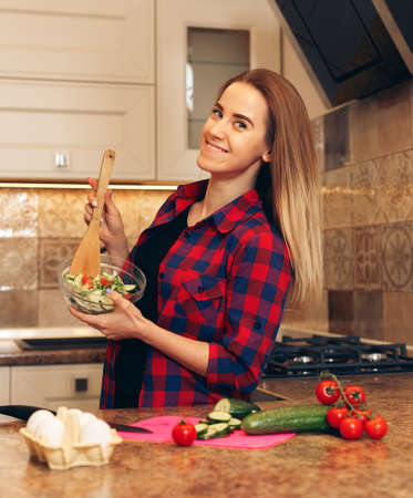 Starting day with healthy food. Beautiful young mixed race woman eating salad and looking away while standing in kitchen at home.の写真素材