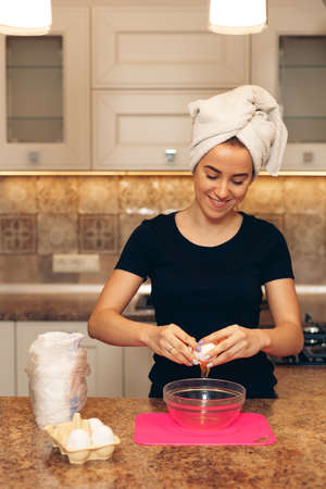 Young woman with a towel on her head preparing breakfast. Happy girl breaking eggs while cooking in the kitchen.の写真素材