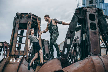 Man with a beard helps a girl to climb on rusty elements. Couple of young people in love on city of urban background.の写真素材