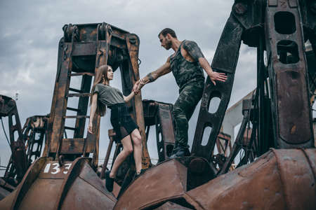 Young man and woman in passion emotions of the street on city of urban background. Man with a beard helps a girl to climb on rusty elementsの写真素材