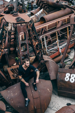 Brutal man with a beard sits on a metal rusty ladle of an abandoned factory. Lifestyle fashion...
