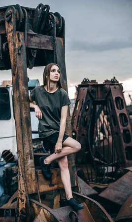 Beautiful young woman sits on a metal rusty detail, an abandoned factory. Cute young stylish woman over urban background.の写真素材