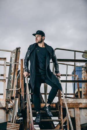 Young attractive man in urban background stands on a metal rusty staircase. Metal rusty ladle of an abandoned factory.の写真素材