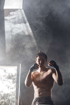 Strong boxer in the rays of the sun and smoke trains in the gym. Handsome guy is training without a T-shirt and hands with boxing wraps.の写真素材