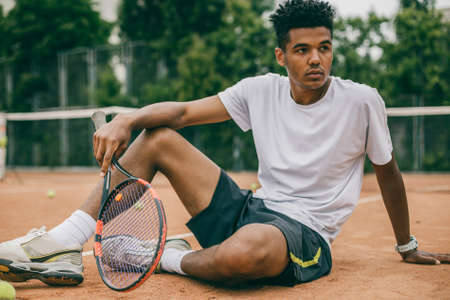 African man and short break during the tennis game. Young man in sports uniform sits on the ground of a tennis court.の写真素材