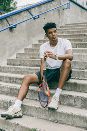 Handsome black guy in polo sits on concrete steps. Tennis player holds racket and ball.の写真素材