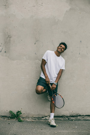 Cheerful young man in sports clothes carrying tennis racket while standing against grey background.の写真素材