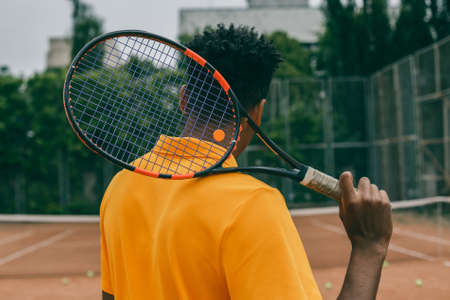 Back view portrait of a man playing in tennis outdoors. Young african man holds a tennis racket on his shoulder.の写真素材