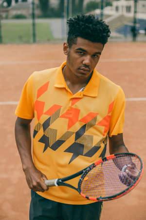 Cheerful young man in orange polo shirt holding tennis racket while standing on tennis court. Great day to playの写真素材