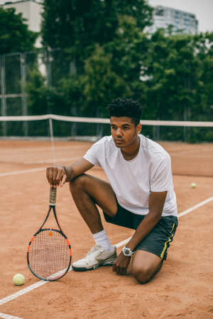 Young man hanging out in the tennis courts sitting on ground. African man resting and chilling on the break time.の写真素材