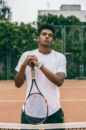Confident young man holding tennis racket and looking at camera while standing on the court.の写真素材