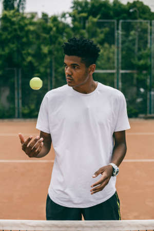 Handsome black guy in polo throws up a tennis ball on the court. Portrait of serious african man looking away.の写真素材