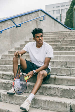 Portrait of serious african man sits on concrete steps. A beautiful black guy in polo and tennis equipment.の写真素材