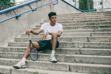 Confident young man in sports clothes on concrete steps. Man holding tennis racket and looking away while sits against grey background.の写真素材