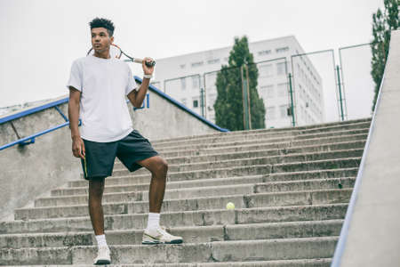 Side view of serious young man in polo shirt carrying tennis racket on shoulder and looking away while standing on concrete steps.の写真素材