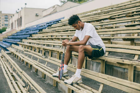 Professional tennis player resting from tennis match. African man sitting on a bench holding his head down.の写真素材