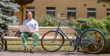 Businessman working on laptop sitting on a bench, next to a bicycle. Handsome man in casual clothes working outdoorsの写真素材