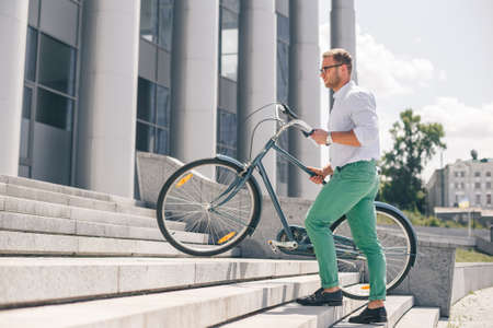 Young stylish businessman carrying bicycle up steps. Handsome man and his bike, concept bike go to work.の写真素材