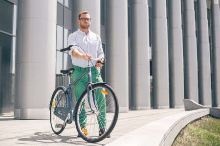 Cool young bearded hipster with bicycle on the city street. Smiling casual businessman having a break. He is standing in front of the building next to bike and holding laptop.の写真素材