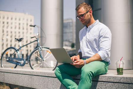 Busy happy man working on laptop outside corporate office. Next to bike. Concept of modern green lifestyleの写真素材