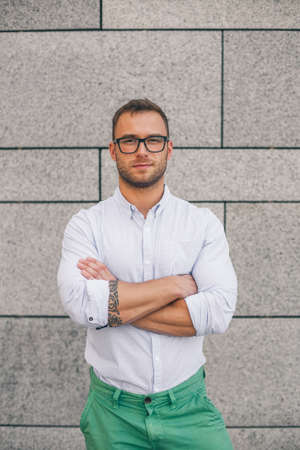 Cool young bearded hipster man wearing shirt, keeping arms crossed and looking at camera while standing against concrete wall background.の写真素材