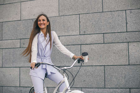 Beautiful young woman on bicycle with coffee. Businesswoman smiling and cycling to work.の写真素材