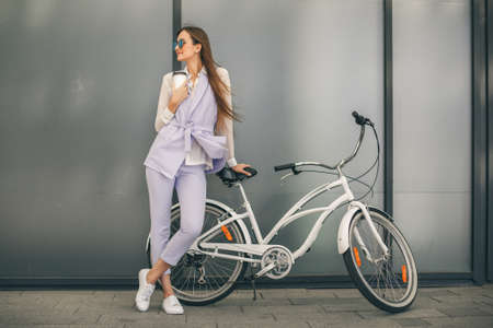Relaxing on a coffee break. Beautiful young woman holding a cup of coffee and smiling while standing on her vintage bicycle near the wall.の写真素材