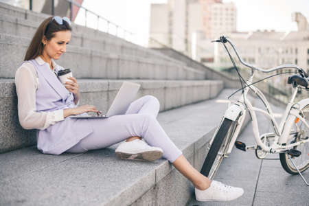 Beautiful woman office worker sitting on the steps and working on a laptop, next to the bike. Girl at lunch break against the backdrop of urban city.の写真素材