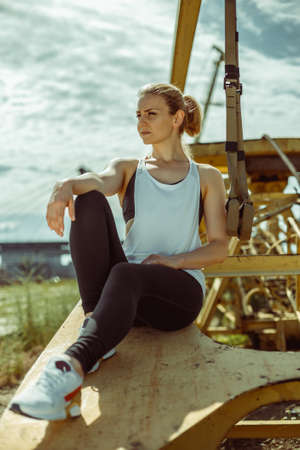 Positive sporty female exercising with trx strips. Beautiful woman in sports uniform sits on metal details.の写真素材
