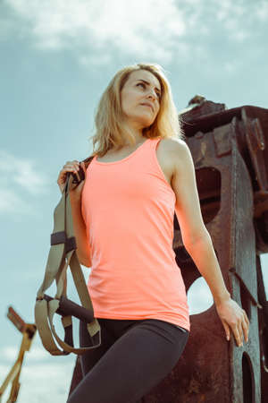 Blonde fitness woman posing with trx fitness straps on metal rusty surface. Beautiful muscular woman doing exercise with trx system.の写真素材