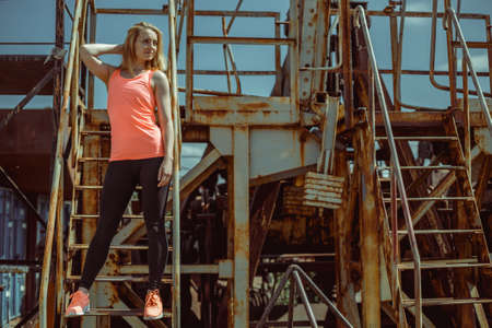 Beautiful young woman in an orange T-shirt is standing on rusty steps. Sport in urban atmosphere city.の写真素材