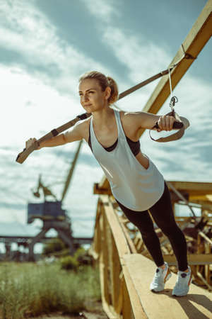 Young attractive woman doing upper body exercise training arms using trx suspension straps outdoor alone. The girl is engaged in sports against the background of metal parts.の写真素材