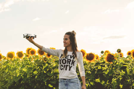Young woman holding on hand flying mini drone outdoors. Beautiful woman is standing near a field of sunflowers at sunset.の写真素材