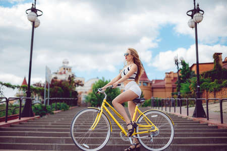 Young beautiful hipster woman in white shorts, summer trend style near beach, riding bicycle. Active fashionable blonde woman riding a vintage bike on urban background at sunset.の写真素材