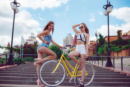 Two cheerful beautiful sexy girls, best friends posing near yellow bike. Dressed in shorts, T-shirts and sunglasses, laugh and screaming smile, crazy, party emotion.の写真素材
