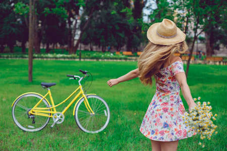 Beautiful blond woman in a straw hat on a lawn with flowers whirls and dances in the park. Bicycle sport and active rest.の写真素材