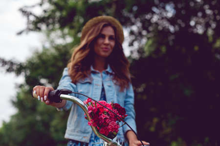 Close-up of a bicycle rudder and blurred background with a young woman in a straw hat in the park. Bouquet of red flowers on the bike.の写真素材