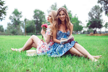 Two beautiful young women sit back to back on the lawn in the parkの写真素材