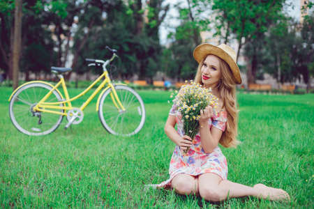 Outdoor portrait of a beautiful blonde woman. Attractive sexy girl in a field with flowers and bike on the background.の写真素材